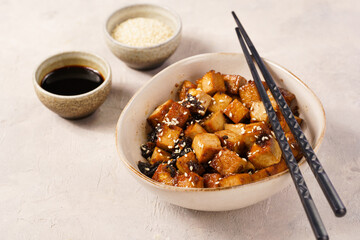 Sticky tofu - vegan soybean protein source in soy sauce and sesame oil marinade roasted on a pan sprinkled with sesame seeds, in a beige colored bowl on a concrete background
