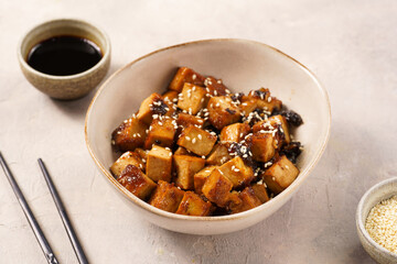 Sticky tofu - vegan soybean protein source in soy sauce and sesame oil marinade roasted on a pan sprinkled with sesame seeds, in a beige colored bowl on a concrete background