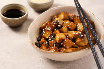 Sticky tofu - vegan soybean protein source in soy sauce and sesame oil marinade roasted on a pan sprinkled with sesame seeds, in a beige colored bowl on a concrete background