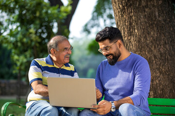 Young indian man showing bank card and laptop how to uses to his father at park