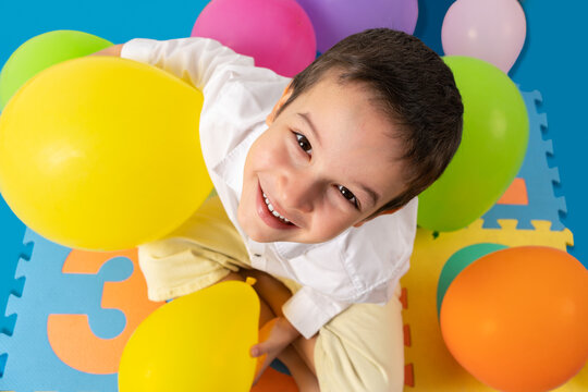 Portrait of an adorable little boy sitting on the floor and playing with among colorful balloons at home