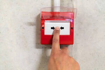 Close up of male hand pointing at red fire alarm switch on concrete wall in office building. Industrial fire warning system equipment for emergency. - obrazy, fototapety, plakaty
