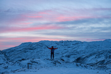 Naklejka na ściany i meble A hiker stands on a rock and looks towards the mountain peaks with outstretched arms in the winter at sunset
