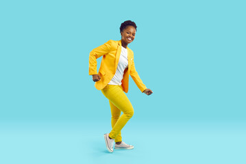 Fototapeta na wymiar Full body shot of happy cheerful joyful beautiful young African American woman in bright yellow suit and white T shirt dancing isolated on blue color background. Fashion, party, having fun concept