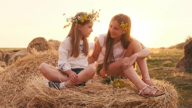 Two pretty sisters wearing flower wreath sitting on hay stack, hugging each other and smiling. Girls spending time in the field in sunset time together. Beautiful female teens out of the city