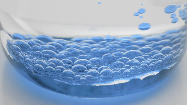 Side view extreme close-up shot of blue transparent bubbles sink to the bottom of beaker with water on grey background | Abstract face care cosmetics with blue tansy oil formulation concept