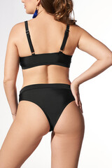 A young lady is wearing a black two-piece swimsuit composed of high-rise bikinis and a shoulder strap bra with a metal ring. The woman is standing with her back to the camera on a light background.