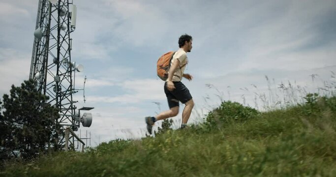 Camera tracking a hiker with an orange backpack running up a hill past the radio tower on a rocky path surrounded with green summer grass.