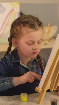 Vertical video. Art school. Kid talent. Creative leisure. Female teacher little girl painting together with paintbrushes on easel in studio.