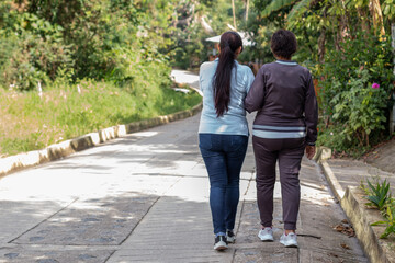 An elderly mother and daughter seen from behind taking a healthy morning walk on a sunny morning.
