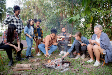 Happy friends group doing recreation with man playing guitar while friends and children grill marshmallow at camping together in holiday, tourist and picnic, party bonfire in vacation.