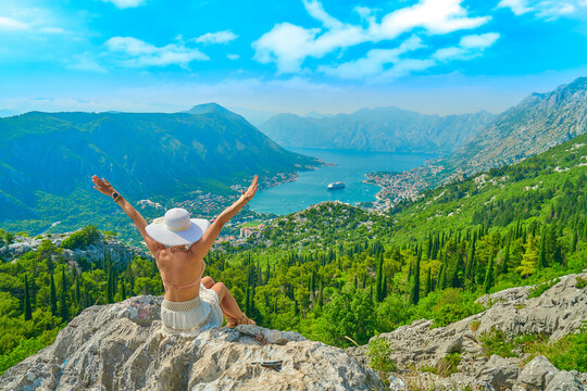 young attractive woman in a hat sitting on a rock with her arms outstretched - in the background a panorama of Kotor Bay with the blue sea, the port and high mountains                              