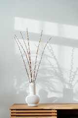 Modern minimalist Scandinavian style interior with blooming branches of the pussy willow in ceramic vase standing on a wooden console with sunlight and shadows on white gray wall. Vertical card
