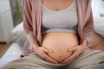 Happy pregnant woman stroking her belly, sitting on bed.