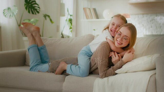 Caucasian Mother In Glasses Lying On Couch Sofa In Home Living Room Little Cute Daughter Kid Child Baby Girl Embrace Hug Cuddle Mom By Neck Piggyback Laughing Together Relaxing Enjoy Hugging Cuddling