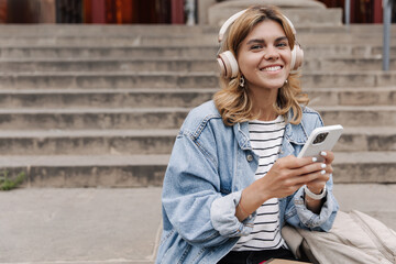 Happy charming woman chatting on phone sitting on the stairs in headphones. Smiling blonde wearing casual clothes outdoors. Concept of lifestyle, technology 