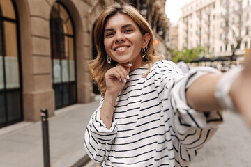 Attractive smiling cute woman reach her hand and look at camera. Happy european lady staying on the street wearing striped shirt. Technology concept