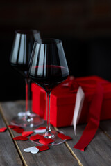 Saint Valentine's Day celebration. Wineglasses with red wine, hearts, gift box, postcard on dark wooden background. Happy holiday . Table decor for festive dinner, romantic atmosphere