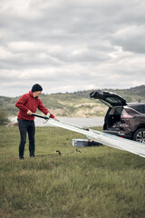 Windsurfer and camper unpacking equipment from a car in nature near the lake shore.