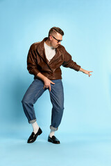 Naklejka premium Self-expression. Portrait of brutal, handsome man in jeans, leather jacket and sunglasses posing, dancing over blue studio background. Concept of emotions, lifestyle
