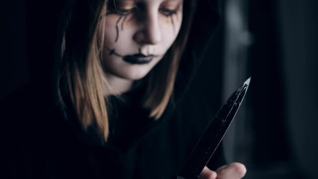 Spooky preteen girl holding knife and looking at blade. Scary child kid in black and Halloween dark makeup