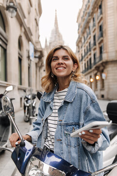 Picture of beautiful european woman in hands phone smiling and looking away. Staying on the street with motobike. Concept of lifestyle, technology 