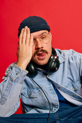 Portrait of man in stylish modern clothes, hat, glasses and headphones posing over red background. Concept of modern fashion, lifestyle, music culture