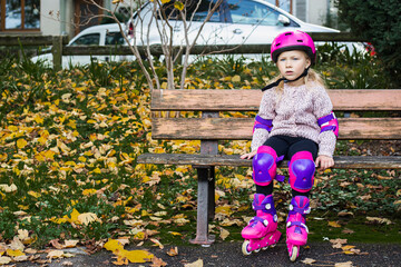 Child girl wearing roller skates in protection sits on a park bench