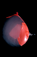 A smoke filled red balloon bursting when shot with an air rifle.  Sound wave visible and pellet exiting.
