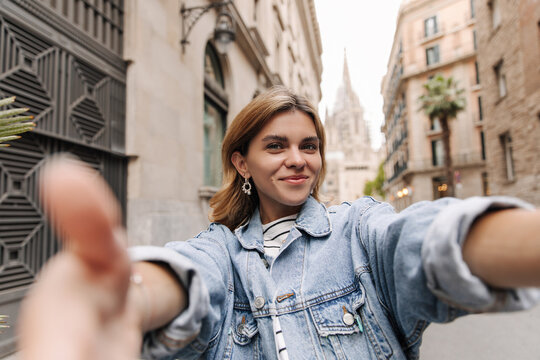 Blonde beautiful woman reach her hands to camera on the street background. Smiling young lady wearing blue jeans jacket. Lifestyle concept