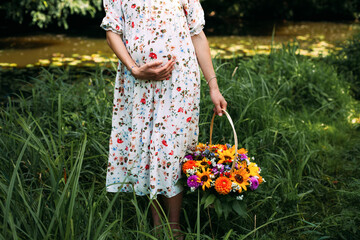 A pregnant girl in a light dress with a bouquet of multi-colored flowers in a basket stands on the grass near the river in spring, holds her stomach with her hand.