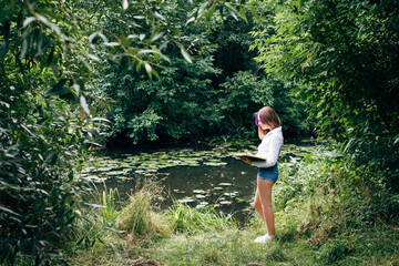 Obraz na płótnie Canvas A young schoolgirl with stylishly dyed hair, with a purple lock of hair, in denim shorts and a white shirt, stands by a picturesque river in the forest and reads a book.