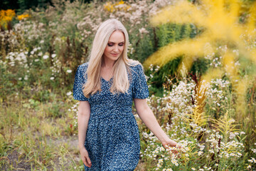 A portrait of a beautiful slender girl with blond long hair holding wildflowers in her hands, in a blue summer dress with a floral pattern, walks along a blooming meadow in summer.