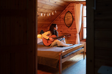 Pensive lonely woman sits on large cozy bed with guitar in hands, plays melody, sadly looks out window, singing favourite song. Rest in country wooden house. Female enjoying playing musical instrument