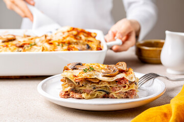 Serving, cooking homemade white lasagna with porcini and champignon mushrooms, onion and meat sausages. Pasta with parmesan cheese and bechamel sauce. Woman hands holding casserole. - obrazy, fototapety, plakaty