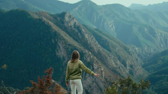 Cinematic woman in traditional sweater hike in mountain. Female hiker reach top of the hill, spread and raise hands in excitement and success. Freedom and joy of travel and outdoor lifestyle.