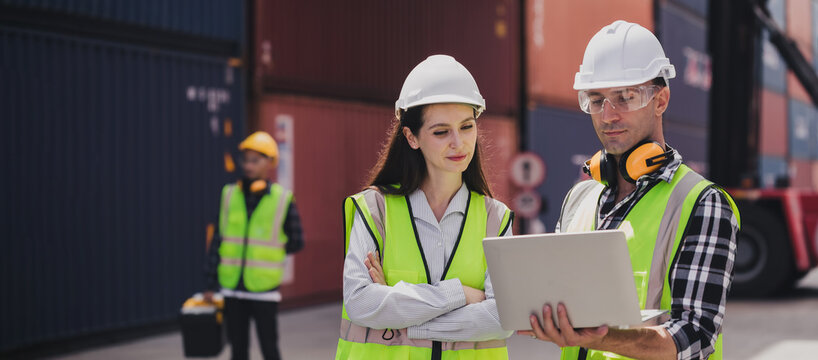 Cheerful Caucasian engineer wearing safety vest standing by shipping container terminal talking discussing information on computer to collaborator. Transportation business. Logistic industrial concept
