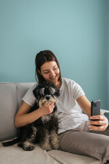 Beautiful caucasian woman takes a selfie with her little black dog miniature schnauzer breed sitting on the couch at home
