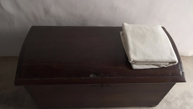 Manos cogiendo toalla blanca doblada sobre un ba&uacute;l de madera antiguo.