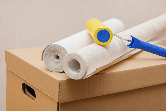 Two rolls of wallpaper, a yellow roller lie on a cardboard box. Repair in the house, moving to a new apartment