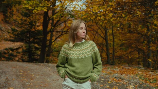 Young woman in traditional sweater in fall forest. Girl look at the camera and smile, calm and relax moment while walking on a warm day in Europe. 