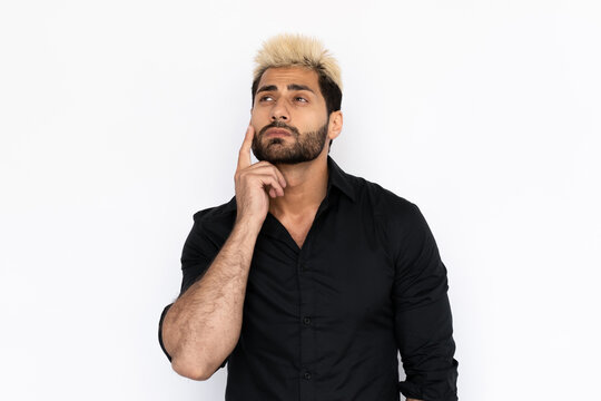 Pensive young man holding finger on his cheek. Male Caucasian model with brown eyes, ombre painted hair and beard in black shirt touching cheek with finger thinking. Advertisement, thought concept