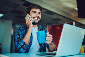 Cheerful man talking on smartphone at table with coffee cup in hand