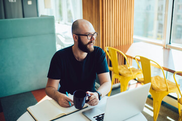 Happy man with coffee cup in hand and laptop at table sitting in cafe