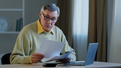 Worried puzzled old caucasian grey-haired businessman 70s male sad man sitting at table with paperwork problems with documents mistake finance budget checks data has conference video call