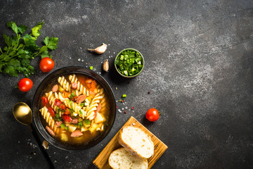 Vegetable soup with pasta and sausages. Fresh soup with zucchini, carrot, paprika, tomatoes and fried sausages. Top view at black stone table.