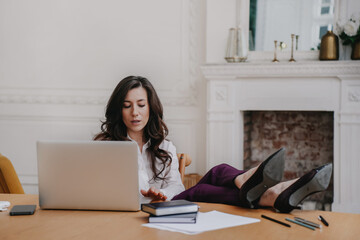 Puzzled brunette young woman in white shirt, dark pants  sitting at desk with laptop puts legs on...
