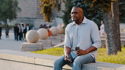 Obraz premium African American ethnic middle-aged man mature senior businessman male sitting outdoors in city with coffee tea and phone waiting for date business meeting looking for friend relaxing on work break