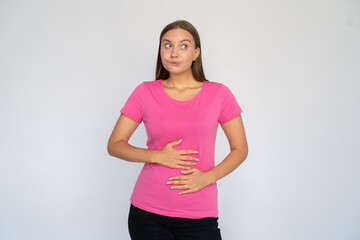 Frustrated young woman with hands on stomach. Portrait of upset Caucasian female model with brown hair in pink T-shirt looking aside, biting lip, suffering from stomach pain. Pain, sickness concept