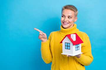 Fototapeta na wymiar Photo of attractive nice short blonde hair girl wear knitted jumper finger point look empty space hold little house isolated on blue color background
